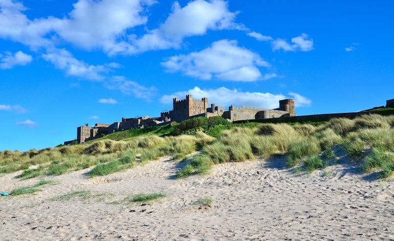 View of Bamburgh Beach with beaches, sandy dunes and the historic Bamburgh Castle in the background, under a bright blue sky with scattered clouds.