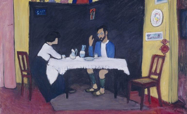 The painting "Kandinsky and Erma Bossi at the Table" by Gabriele Münter, 1912, depicts two figures sitting at a table in a domestic interior. The man, likely Kandinsky, is seated on the right with a blue jacket, while a woman, likely Erma Bossi, is seated on the left. The background features a dark wall with shelves and various objects, and the room is adorned with simple furniture and decorations, creating a cozy, intimate setting. A good example of Expressionism.