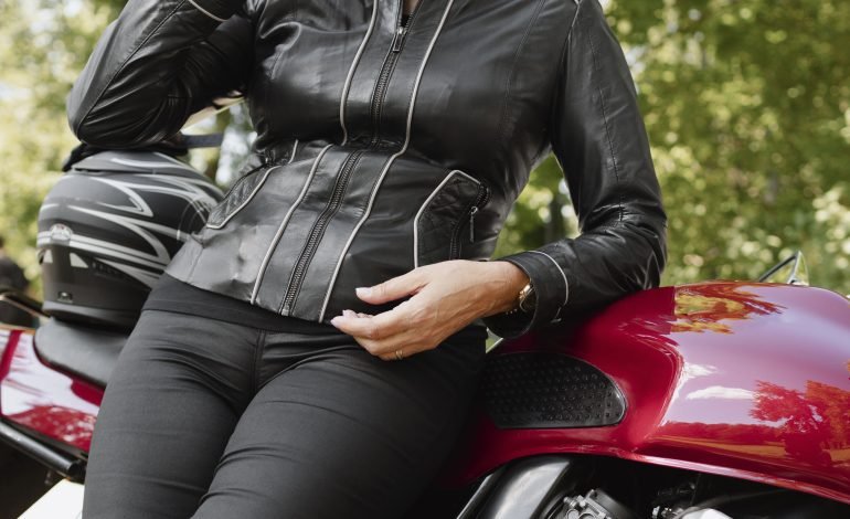 Confident ageing woman in a leather jacket, sitting on a red motorcycle, adjusting her sunglasses with a smile, set against a backdrop of lush green trees.