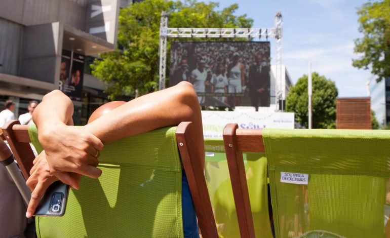 People relax in green deckchairs in the sun and watch a sporting fixture on a large outdoor screen. This summer, Hammersmith residents can watch the Paris 2024 Olympics on the big screen in London.