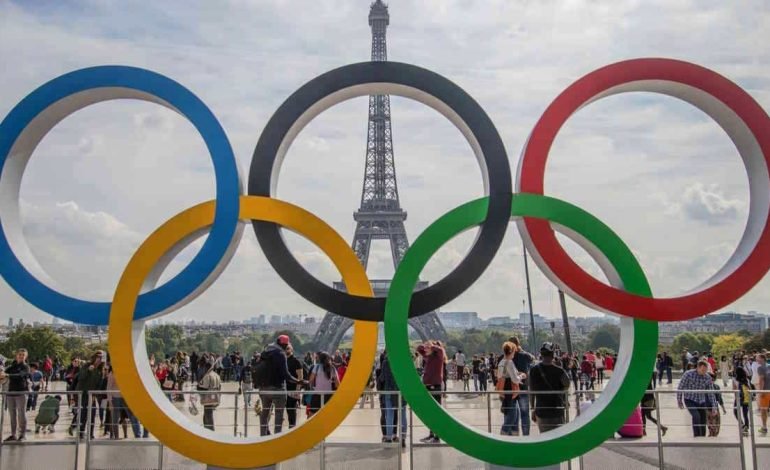 Olympic rings in front of the Eiffel Tower, Paris, with crowds gathered, celebrating the start of the Paris Olympics, featuring football action.