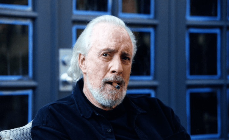 Portrait of Robert Towne, an elderly man with white hair and a beard, sitting and smoking a cigar, wearing a dark shirt with a contemplative expression.