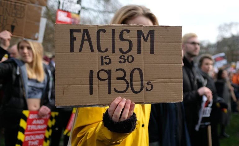 A protester holding a sign saying "Facism is so 1930s" and " in a city centre with police presence in the background.