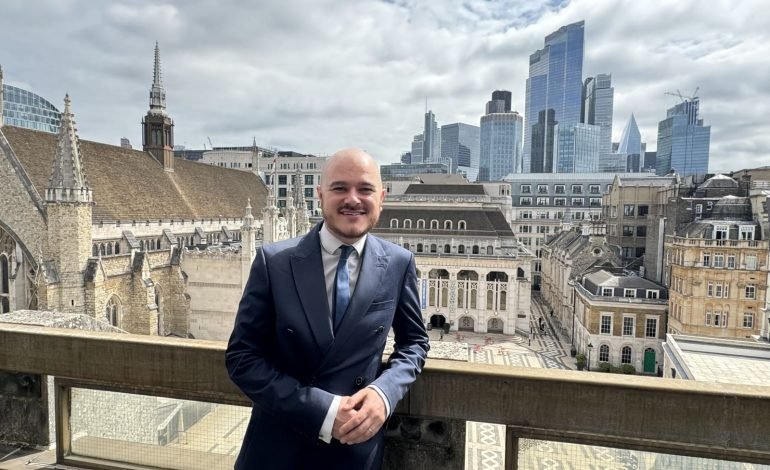 Greg Moore, newly appointed Deputy Town Clerk of the City of London Corporation, standing on a balcony with the Guildhall and the modern City of London skyline in the background.