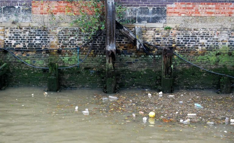 Polluted riverbank with visible litter and dilapidated wooden structures, showcasing environmental degradation in urban waterways, underscoring the urgency of Ofwat and the recent fines on major water companies for sewage mismanagement.