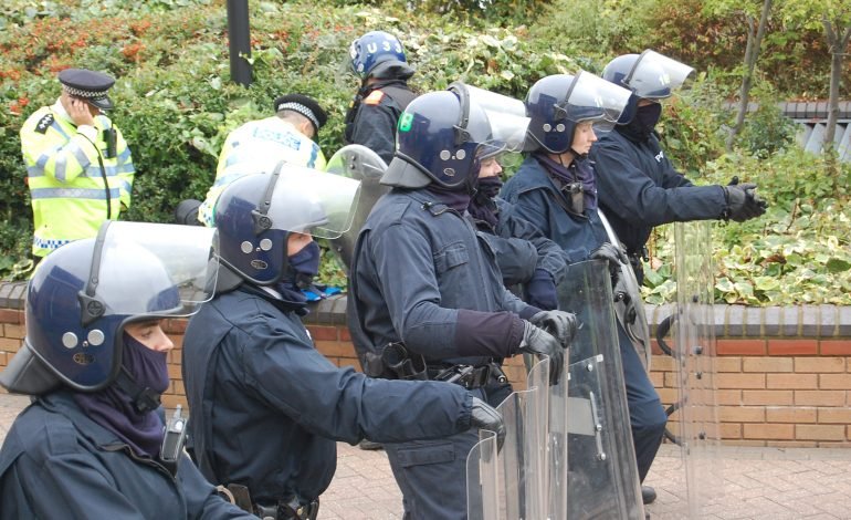 Riot police in protective gear standing in formation during UK unrest.