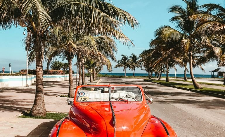 Classic red vintage car parked under palm trees near a beach in Cuba.