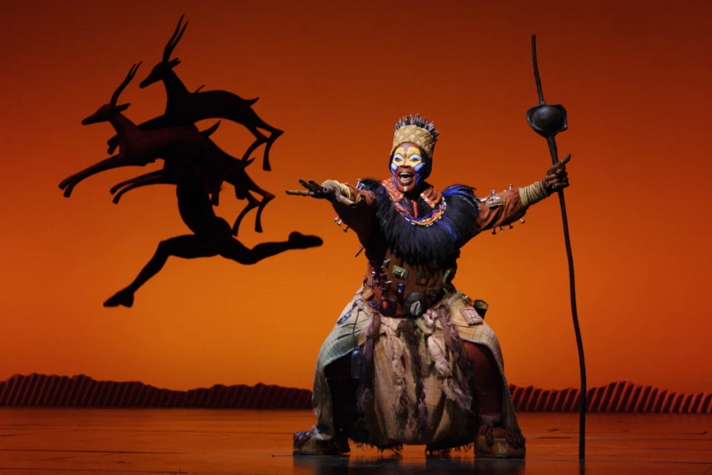 Rafiki commands the stage in The Lion King at the Lyceum Theatre, London, as silhouetted antelope race across the rising sun backdrop. Photo Johan Persson