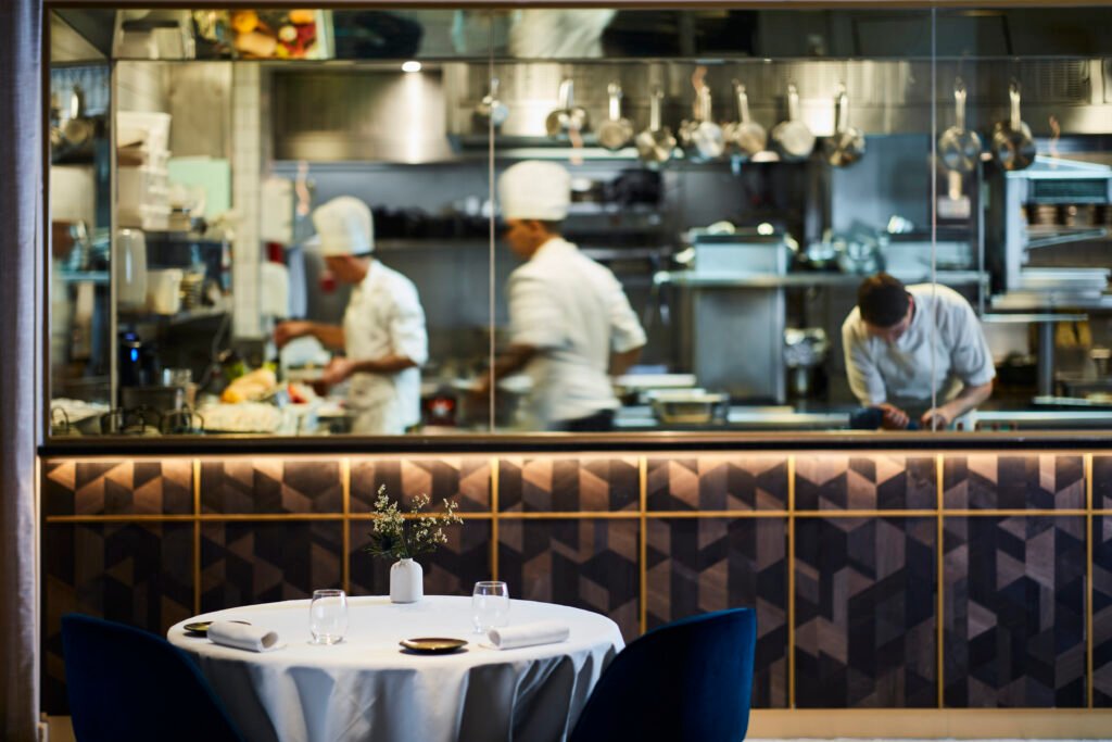 Image 2 Open kitchen at CORD by Le Cordon Bleu with chefs preparing dishes behind a table set for two.