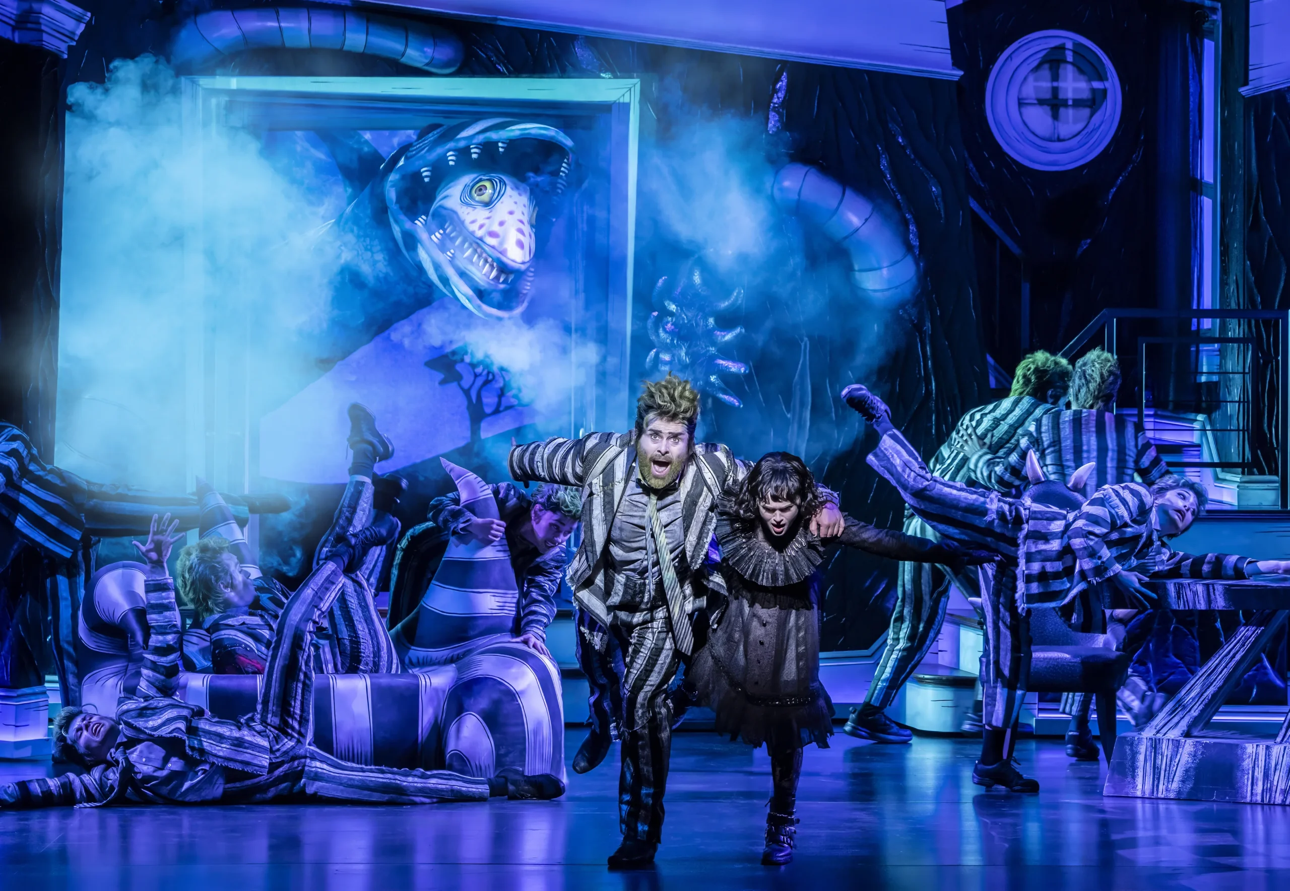 David Fynn as Beetlejuice and Hannah Nordberg as Lydia Deetz, surrounded by the company in a stage scene from Beetlejuice the Musical