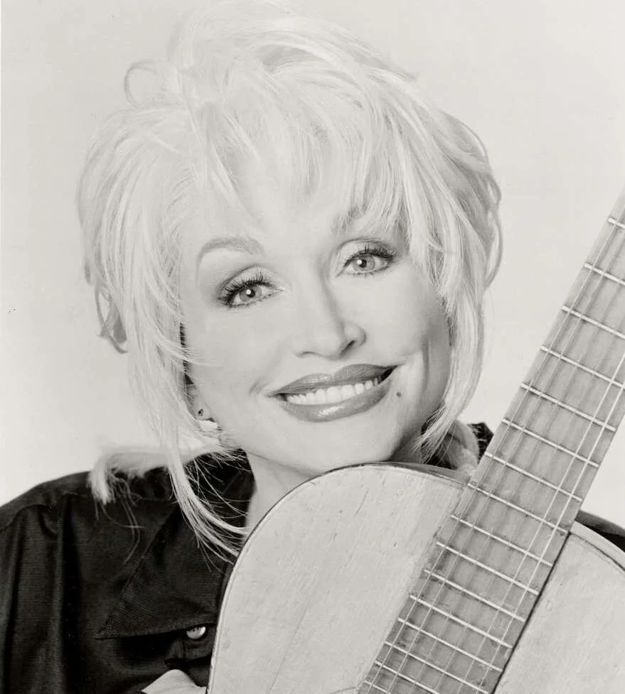 Dolly Parton smiles in a black and white portrait holding a guitar, star of Dolly Parton musicals
