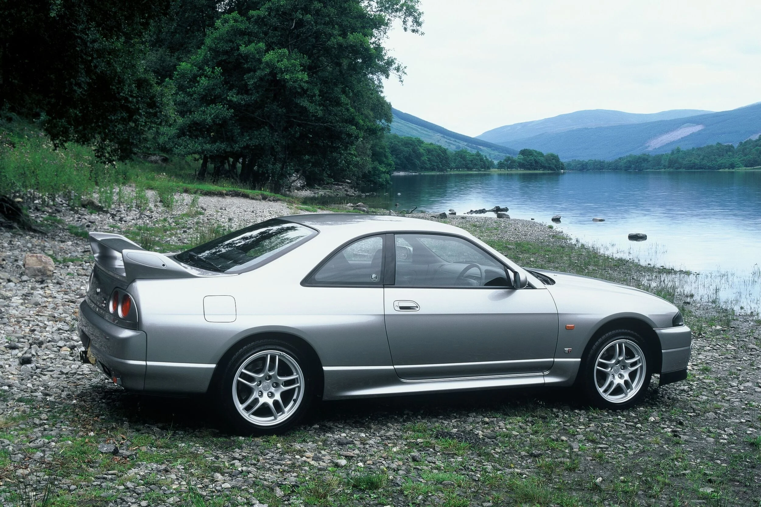 a stunning silver Nissan Skyline R33 GTR besides a lake