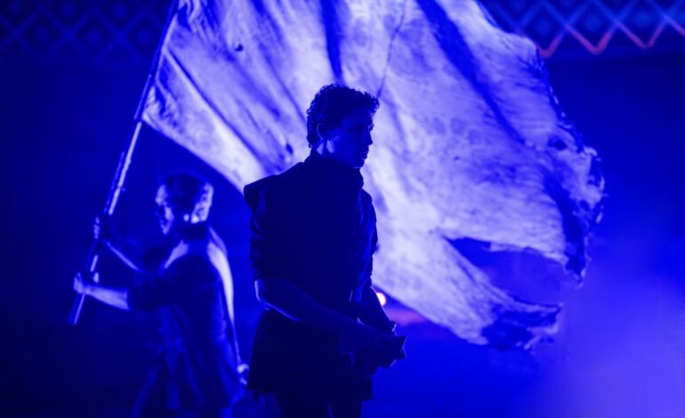 Martin Guerre cast members under blue light with a war banner at the Old Vic